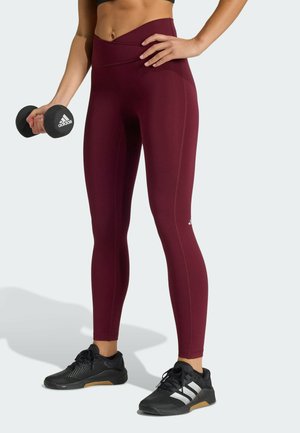 Donkerrode legging met hoge taille en gestructureerde stof. Het model houdt een zwarte halter in één hand en draagt zwarte sportschoenen met witte strepen.