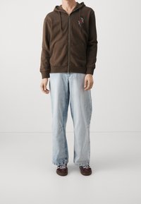 Uomo che indossa una felpa marrone con zip e design floreale, jeans larghi blu chiaro e sneaker marroni scuri con lacci bianchi, in piedi contro uno sfondo uniforme.