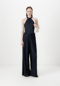 Navy pleated jumpsuit with halter neck, wide legs, and a cinched waist. Model holds a small white handbag with a textured surface.