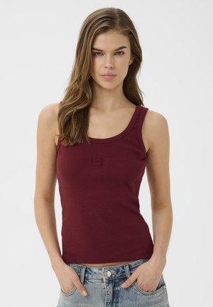 Młoda kobieta o długich brązowych włosach, ubrana w dopasowany bordowy top na ramiączkach i niebieskie dżinsy, ręce w kieszeniach, neutralny wyraz twarzy, białe tło.