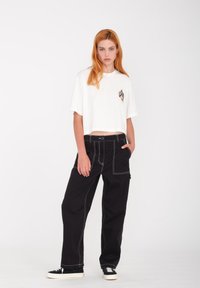Camiseta blanca corta con un gráfico colorido en el pecho, combinada con pantalones negros holgados que tienen costuras blancas y bolsillos.