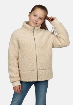 Young girl wearing a beige zip-up fleece jacket and blue jeans, smiling with her right hand touching her hair.