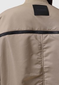 Beige Bomberjacke mit schwarzem Reißverschluss, gesteppten Details und einem rechteckigen Lederpatch auf der Rückseite mit Logo.