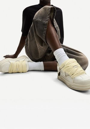 Chunky white sneakers with braided yellow laces, textured materials, and a small metal accent. Paired with gray wide-leg pants and white socks.