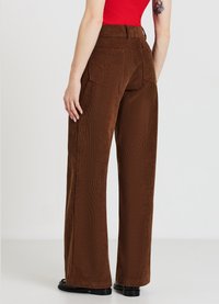 Braune Cordhose mit weitem Bein und strukturierter Oberfläche, ausgestattet mit zwei Gesäßtaschen und hoher Taille. Schwarze Schuhe runden den Look ab.