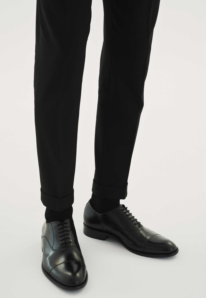 Fekete, sima bőrhatású bőrcipő, lekerekített orral és díszítő varrással. Fekete nadrággal viselve, amely felhajtott szegélyű.