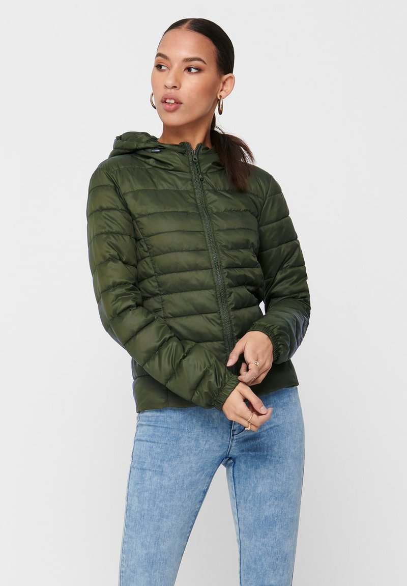 Polstret olivengrønn jakke med glidelås og hette, med en quiltet design og elastiske mansjetter. Brukt over lyseblå jeans.