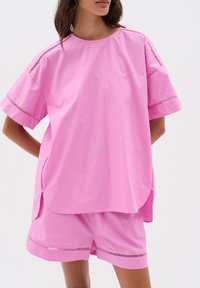 Růžový bavlněný oversized top s krátkými rukávy, s bočními rozparky a dekoračním prošíváním podél švů, doplněný o odpovídající šortky.