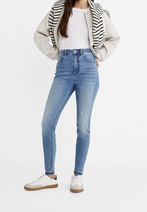Femme debout, mains sur les hanches, portant un jean skinny taille haute bleu clair, des baskets blanches, un haut blanc et un pull rayé posé sur les épaules.