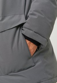 Grå polstret jakke tæt på, der viser et tekstureret stof, synlig lomme søm, elastisk manchet og et slankt, moderne design.