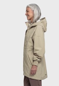 Lächelnde ältere Frau mit grauem Haar, die einen beigefarbenen Regenmantel mit Kapuze und braune Hosen trägt, im Profil vor einem weißen Hintergrund stehend.