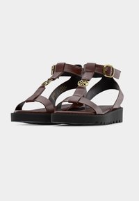 Καφέ δερμάτινα σανδάλια με σόλα πλατφόρμα, διαθέτουν σχεδιασμό με λουράκι Τ, αγκράφα στον αστράγαλο και διακοσμητικά στοιχεία σε χρυσό τόνο. Υπόδημα με υφή στην μαύρη εξωτερική σόλα.