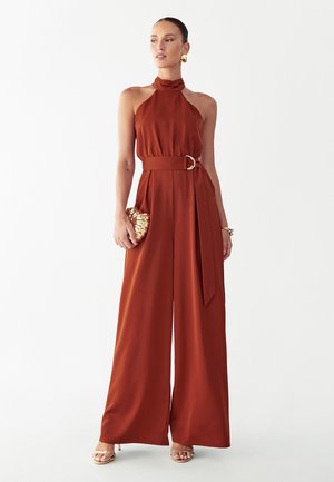 Femme portant une combinaison à col nageur de couleur rouille avec jambes larges, taille ceinturée, boucles d'oreilles dorées, bracelet, pochette en forme de coquillage et talons dorés.