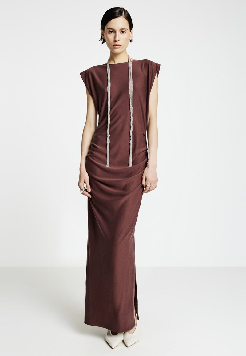 Robe longue de couleur brun chocolat avec manches courtes structurées, tissu drapé, fente latérale et un collier décoratif à pampille argentée.