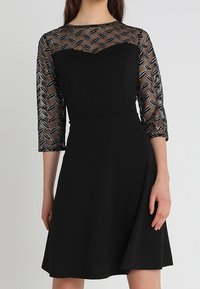 Svart klänning med trekvartslånga ärmar, med en genomskinlig topp med ett geometriskt mönster och en figurnära bodice som vidgar sig vid midjan.