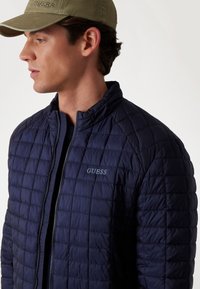 Jachetă matlasată de culoare bleumarin cu guler în picioare, fermoar frontal și logo-ul "GUESS". Prezintă o textură fină și un model de coaseri pătrat. Asortată cu o șapcă oliv.