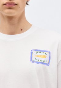 Νεαρό άτομο που φοράει λευκό πουκάμισο με πολύχρωμο γραφικό κονσέρβας σαρδέλας και κείμενο «Original Sardines» στο αριστερό στήθος.