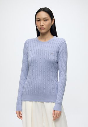Young woman with straight black hair wears a light blue cable-knit sweater and white pants, standing against a plain white background.