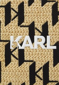 Vävd halmtextur med ett svart geometriskt mönster; har den vita texten "KARL" med en utskärning av ett ansikte i bokstaven "K."