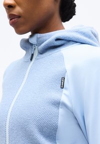 Κοντινό πλάνο ενός ατόμου που φοράει ανοιχτό μπλε ζακέτα με υφή και φερμουάρ, με μια μικρή μαύρη ετικέτα στο στήθος.