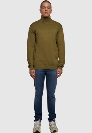 Jeune homme debout portant un pull à col roulé vert olive, un jean bleu et des baskets beiges sur un fond gris uni.