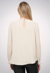 Blusa beige a maniche lunghe con tessuto texturizzato, dotata di un'apertura a chiave sul retro e polsini elasticizzati. Finitura liscia con una vestibilità morbida.
