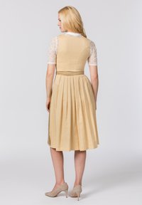 Ein senfgelbes Kleid aus gemustertem Stoff mit tailliertem Schnitt und plissiertem Rock. Es hat einen transparenten Spitzenärmel und beigefarbene Absätze.