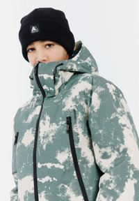 Groene en witte gestreepte winterjas met hoge kraag, zwarte ritsen en logo detail, gecombineerd met een zwarte gebreide beanie.