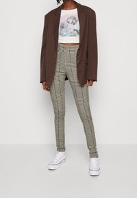 Blazer marrón de gran tamaño sobre una camiseta gráfica corta con esculturas; pantalones de cuadros de tiro alto y zapatillas blancas completan el conjunto.