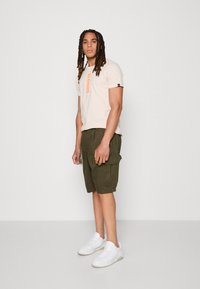 Lyserød t-shirt med orange "ALPHA" logo, sammen med olivengrønne cargo shorts med sidelommer og hvide sneakers.
