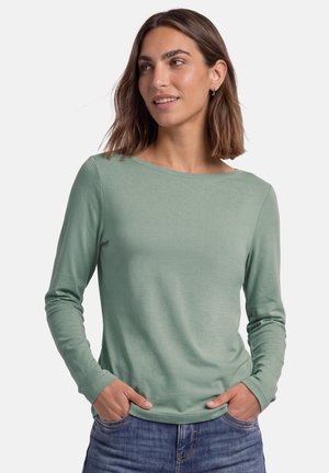 Femme aux cheveux bruns mi-longs, portant une chemise à manches longues vert clair et un jean bleu, les mains dans les poches, regardant légèrement vers la droite.