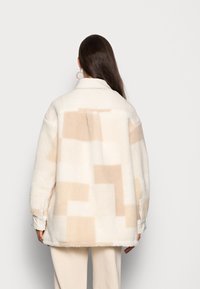 Oversized fleece jasje in crème met abstracte beige blokpatronen. Heeft een kraag, lange mouwen en een relaxte snit.