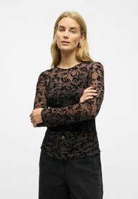 Juodas ilgomis rankovėmis viršus su gėlių nėriniais, turintis apvalią iškirptę ir prigludusią siluetą, derinamas su tamsiomis džinsais.