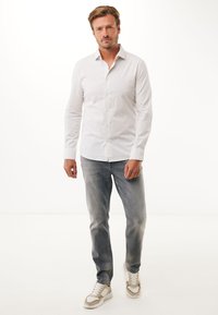 Camisa blanca de botones con finas rayas verticales, combinada con jeans de mezclilla gris claro y zapatillas en tonos neutros. Diseño simple y ajustado.