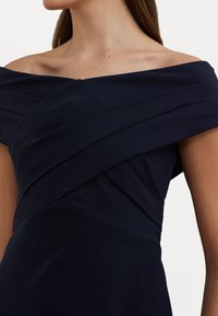 Off-shoulder klänning i marinblått i lätt tyg har diagonala veck över bysten, vilket skapar en strukturerad silhuett.