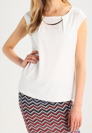 Kvinde iført hvid ærmeløs bluse med plissering ved halsudskæringen og nederdel med flerfarvet zigzag-mønster, hånd på hoften.