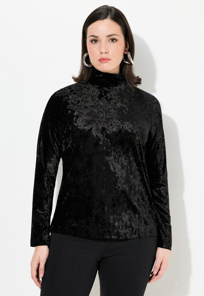 Schwarzes Samtoberteil mit langen Ärmeln und hohem Kragen sowie subtiler Textur, kombiniert mit dunklen Jeans. Runde silberne Ohrringe fügen ein Detail hinzu.