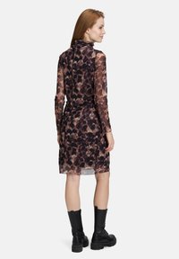 Frau mit roten Haaren, die ein langärmliges Kleid mit braunen und schwarzen Punkten und schwarze Schnürstiefel trägt, steht und schaut seitlich.