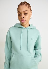 Fleece verde menta claro com bolso frontal, capuz com cordão e pequeno emblema. Tecido suave e liso com punhos canelados.