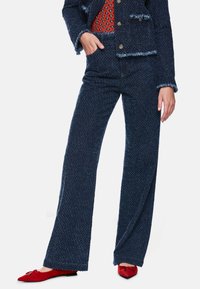 Marineblaues, strukturiertes zweiteiliges Outfit mit kurzem Blazer und ausgestellten Hosen, ergänzt durch rote spitze Ballerinas mit Schleifendetail.