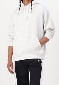 Sudadera gris clara con capucha ajustable, bolsillo frontal y puños acanalados. Combinada con pantalones negros que tienen franjas laterales blancas y un logo.