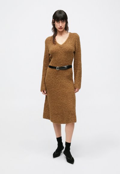 Bruine, pluizige gebreide sweaterjurk met een V-hals en lange mouwen, met een zwarte leren riem opgesnoerd. Draagt zwarte sokken en schoenen met hak.