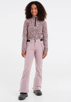 Nuori nainen, jolla on kiharat hiukset ja päällään vaaleanpunainen kuvioitu pitkähihainen paita, vaaleanpunaiset lasketteluhousut ja mustat talvikengät, seisoo ja hymyilee.