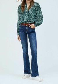 Blommig grön blus med vida ärmar, kombinerad med mörkblå utsvängda jeans och ett brunt bälte. Vita sneakers fulländar stilen.