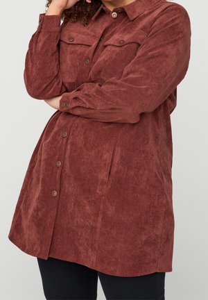 Personne portant une chemise en velours côtelé rouille à manches longues, boutonnée, avec des poches poitrine, bras croisés, sur un fond uni.