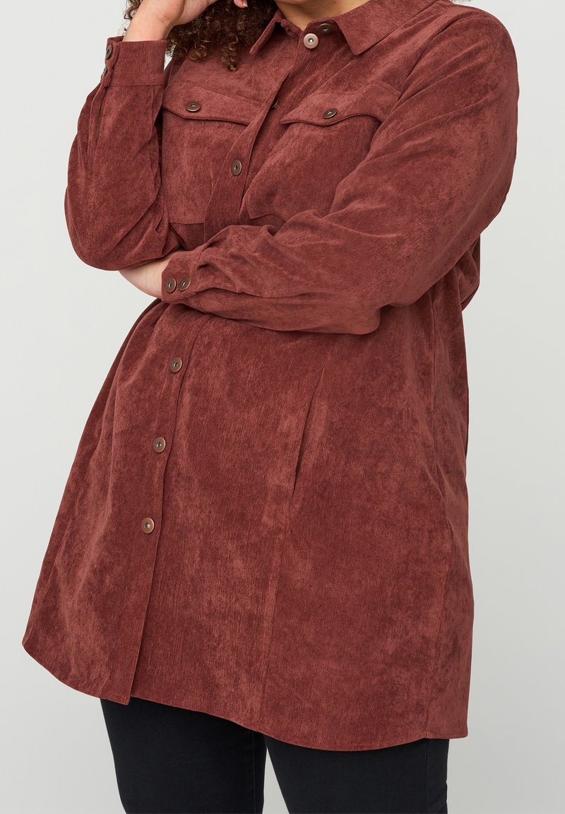 Personne portant une chemise en velours côtelé rouille à manches longues, boutonnée, avec des poches poitrine, bras croisés, sur un fond uni.