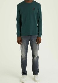 Grön långärmad bomullsskjorta med rund halsringning och svart logotyp. Bärs tillsammans med blekta blå jeans och vita sneakers.