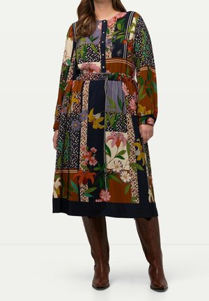 Kvinde iført en langærmet, blomster patchwork midi kjole med knapper foran, kombineret med knæhøje brune læderstøvler.