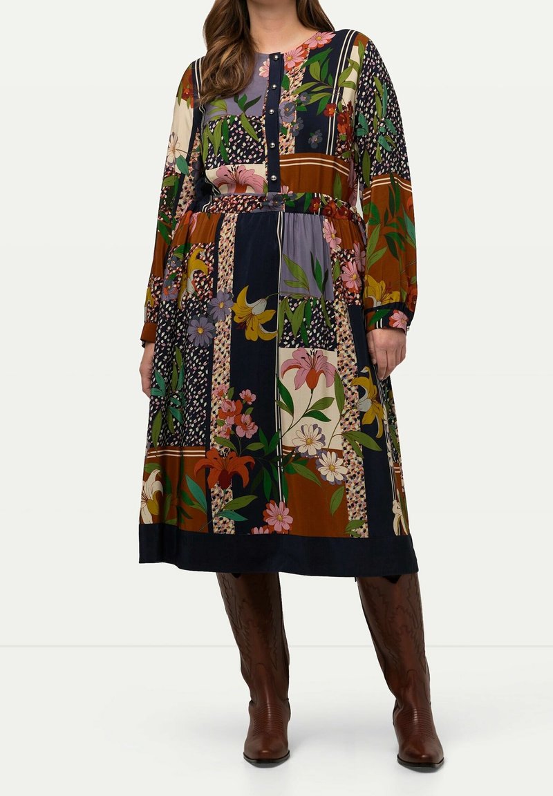 Femme portant une robe midi patchwork florale à manches longues avec des boutons sur le devant, associée à des bottes en cuir marron montant jusqu'aux genoux.