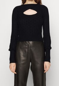 Femme portant un pull noir en maille torsadée avec un col clé et des manches longues, associé à un pantalon en cuir noir taille haute.